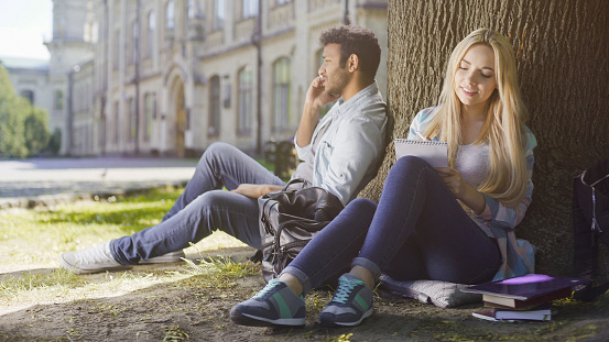Multiracial guy talking on cellphone under tree, girl taking notes in notebook, stock footage