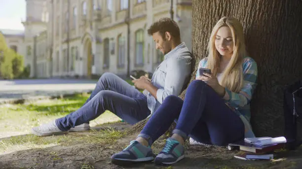 Photo of Multiracial guy and girl sitting under tree using mobile phones smiling, happy