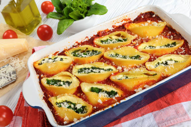 conchiglion pasta stuffed with cheese and greens delicious baked conchiglion pasta stuffed with creamy soft cheese and spinach sprinkled with parmesan cheese in baking dish. grated parmesan cheese on paper, italian recipe, view from above, close-up stuffed shells ricotta cheese stock pictures, royalty-free photos & images