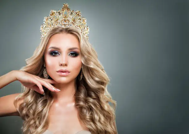 Glamorous Blonde Woman with Golden Crown, Makeup and Wavy Hair. Cute Fashion Model with Diamonds Jewelry, Shiny Curly Hairstyle and Make up