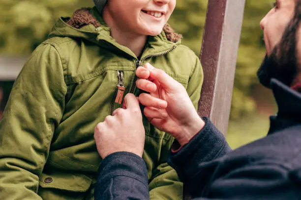 cropped shot of father fastening jacket of cute smiling little son in park