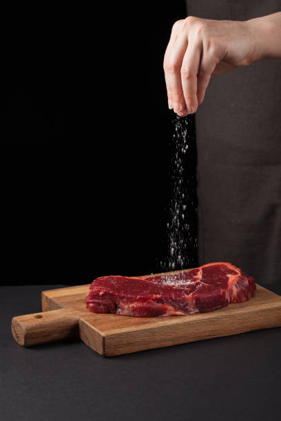 istock Beef steak and salt by chef on the background with free space for text design or logotype menu restaurant. Vertical photo. Food background. Black text area. Copy space 907305644