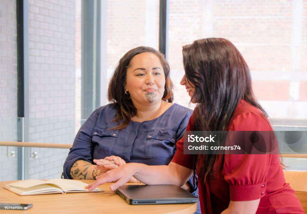 Maori and caucasian business women smiling in meeting, could be coworkers, employees or management. Maori female has moko facial tattoo. Maori and caucasian business women smiling in meeting, could be coworkers, employees or management. Maori female has moko facial tattoo. Photographed in New Zealand, NZ Candid Stock Photo