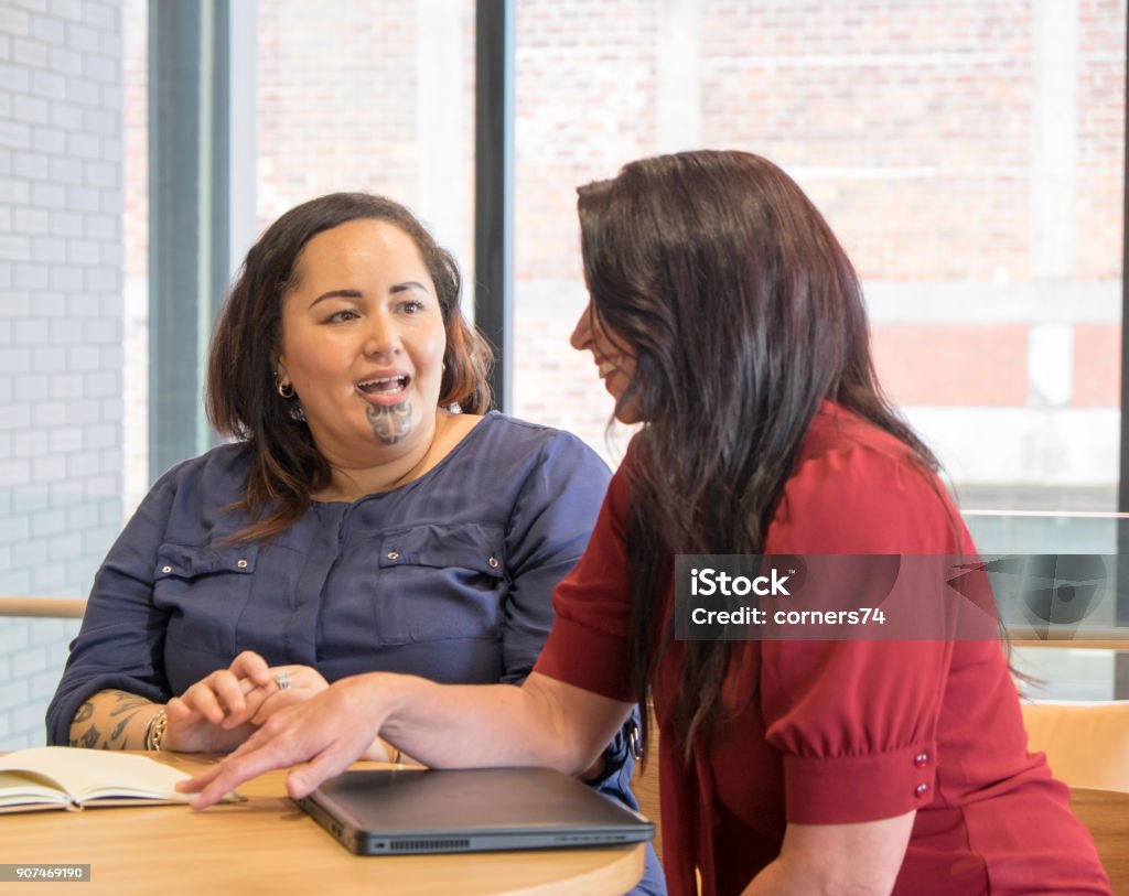 Maoríes y caucásico mujeres hablar en la reunión, podría ser la administración, empleados o compañeros de trabajo. Mujer maorí tiene moko facial tatuaje. - Foto de stock de Diálogo libre de derechos