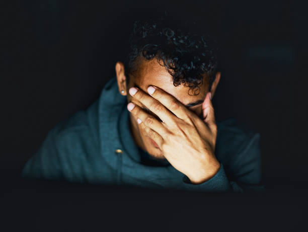 Young man sitting in the dark covers his face A hoodie-wearing young man, seen sitting in the dark, holds his hand over his face, exhausted or miserable. head in hands stock pictures, royalty-free photos & images