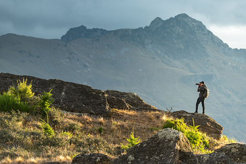 Young male photographer taking photo in photographing posture with mountain scenery during golden hour sunset in Queenstown, South Island, New Zealand. Travel and photography concept