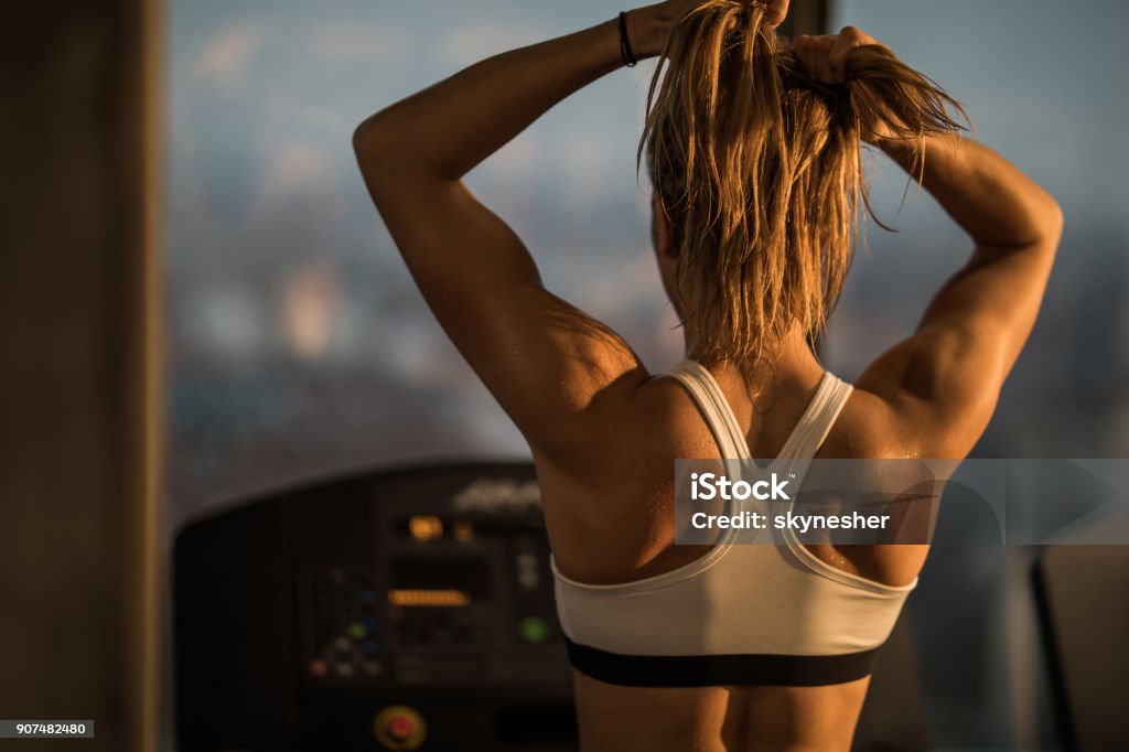 Back view of athletic woman making ponytail before sports training in a gym. Rear view of muscular build woman tightening her ponytail before exercising on treadmill in a health club. Muscular Build Stock Photo
