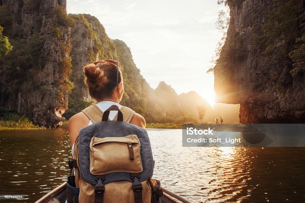 woman with backpack traveling by boat enjoying sunset among of mountains. Woman with backpack swims on boat among karst mountains to meet her friends. Tam Coc,  North of Vietnam. Travel Stock Photo