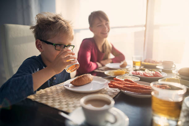 Mother and kids eating breakfast.  There are dry sausages, lemon, bread, butter, coffee, juice, and ham on the table.\n