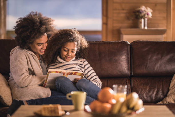 relajado la madre afroamericana e hija leyendo un libro en el sofá. - acogedor fotografías e imágenes de stock