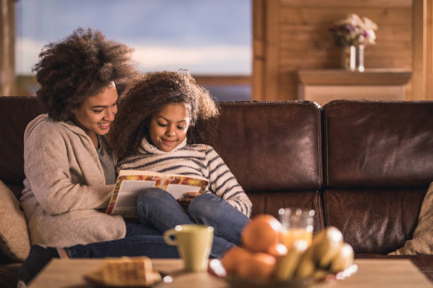 Relaxed African American mother and daughter reading a book on sofa. Happy black mother and daughter relaxing on sofa at home and reading a book. cozy stock pictures, royalty-free photos & images