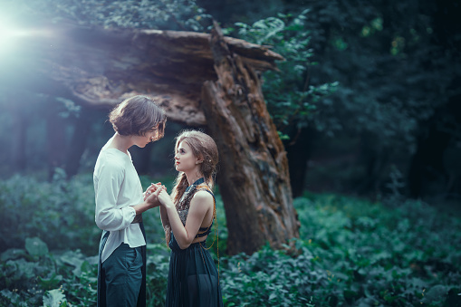 Young couple of elves in love in magical forest agaist the broken tree outdoor on nature at night. Fairy tale love, relationship and magic people concept.