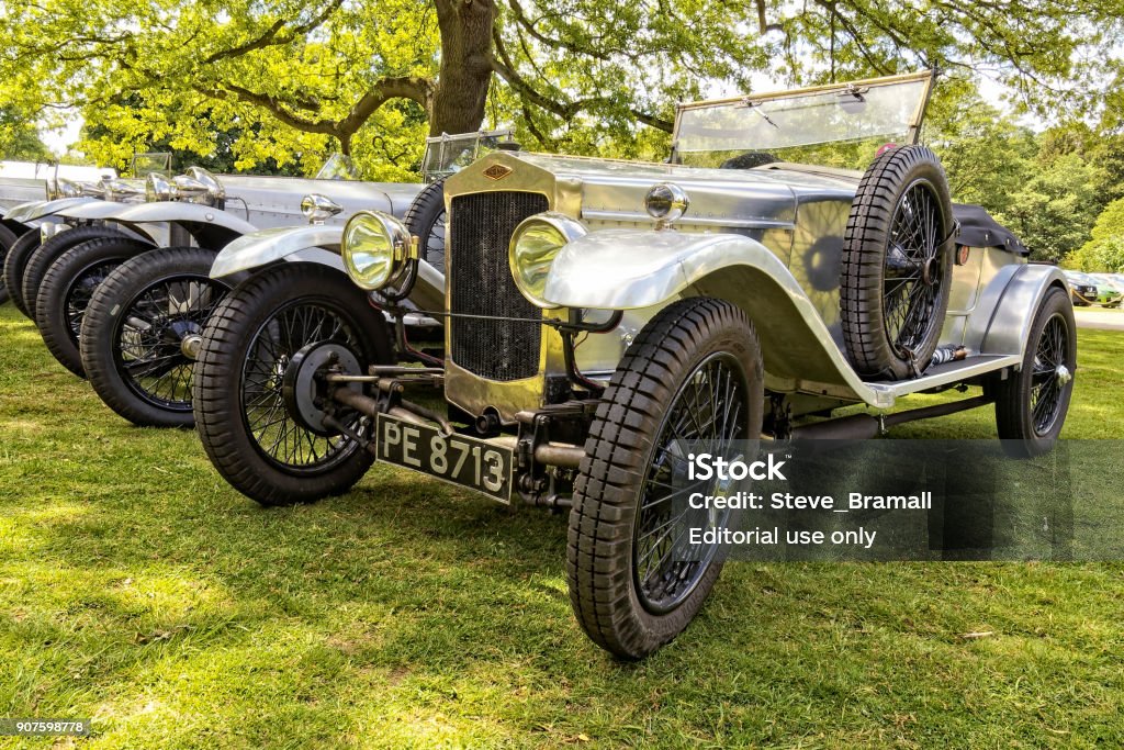 Line of silver Frazer-Nash cars Leicester, England - June 6th 2015: A group of vintage silver Frazer-Nash convertible sports cars, all with the soft top removed, parked in a line under trees on short grass. The closest car has two doors, a brass and black coloured radiator, large headlights, spare wheel strapped to the side of the car and black wire wheels, A front three-quarter view image. Antique Stock Photo