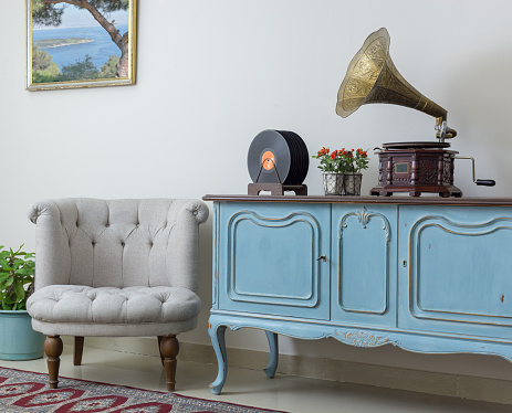 Retro off white armchair, vintage wooden light blue sideboard, old phonograph (gramophone) and vinyl records on background of beige wall, tiled porcelain floor, and red carpet