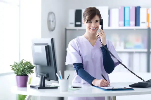 Young practitioner doctor working at the clinic reception desk, she is answering phone calls and scheduling appointments