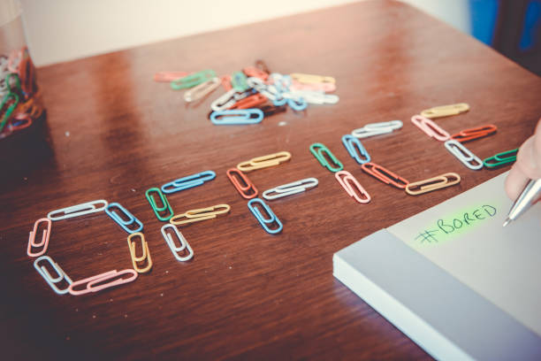A conceptual image representing people being bored at their place of work.  A woman hand scribbles #Bored on a notepad, while she has formed the word ‘office’ from paper clips.  Image taken in Ko Lanta, Krabi, Thailand.
