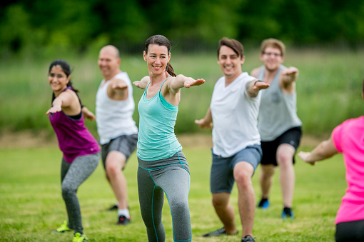 A multi-ethnic group of men and women are outdoors. They are doing a boot camp together. They are doing yoga now, and stretching their arms out.