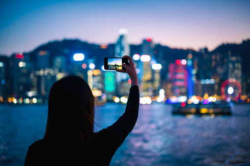 Young woman using a smartphone to take a picture of the Victoria Harbour night view.