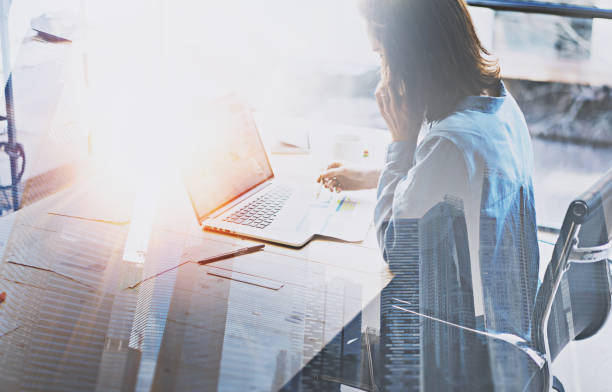 istock Businesswoman working process at modern office.Account manager work wooden table with paper documents and mobile laptop.Double exposure,skyscraper building blurred background.Horizontal.Flares effect. 907793502