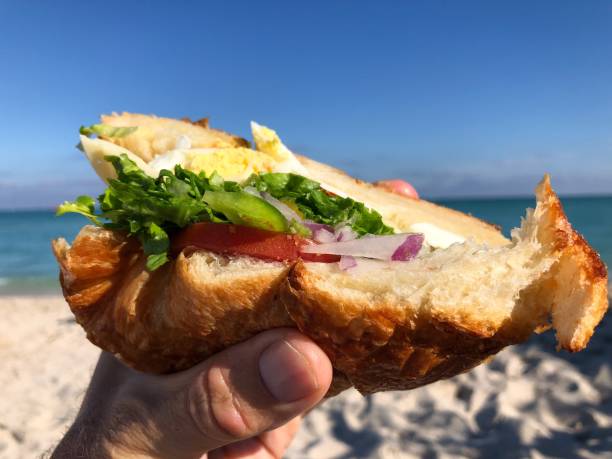istock Hand Holding Croissant Sandwich on the Beach 907805582