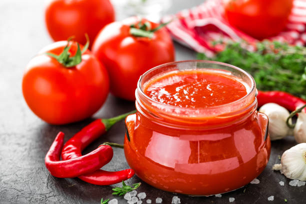 istock Tomato paste, puree in glass jar and fresh tomatos on dark background. Hot vegetable sauce with chili pepper ant tomatoes 907848012