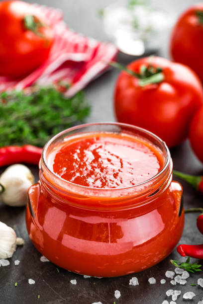 istock Tomato paste, puree in glass jar and fresh tomatos on dark background. Hot vegetable sauce with chili pepper ant tomatoes 907848014