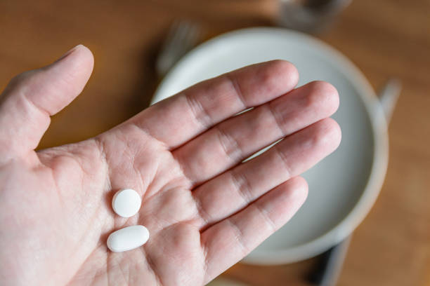вид крупным планом белой мужской руки, держащей две белые таблетки на ладони над �размытым фоном, показывающим обеденный стол с тарелкой, ст� - ibuprofen стоковые фото и изображения