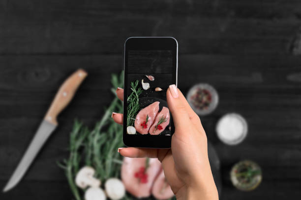 female hand with a smartphone makes a photo raw chicken breast on black background, with fresh mushrooms, garlic and herbs - garlic chicken breast raw chicken imagens e fotografias de stock