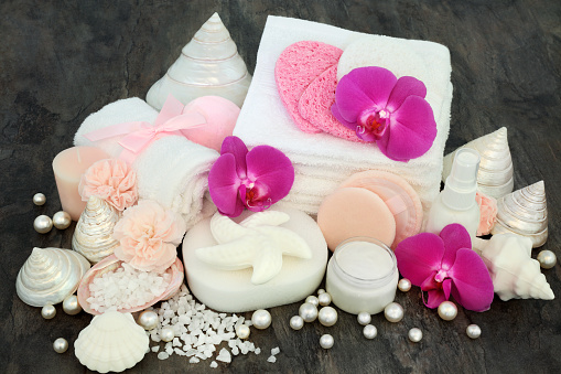 Cleansing spa and ex foliation beauty treatment with orchid and carnation flowers, moisturising cream, ex foliating salt, shell  shaped soaps, sponges, wash cloths, body lotion with decorative seashells and pearls on marble background.
