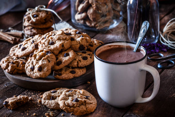 Homemade chocolate chip cookies and hot chocolate mug Hot chocolate mug shot on rustic wooden table. Some baked cookies are on a plate beside the mug. A cokie jar is visible at background. Predominant color is brown. Low key DSRL studio photo taken with Canon EOS 5D Mk II and Canon EF 100mm f/2.8L Macro IS USM hot chocolate stock pictures, royalty-free photos & images