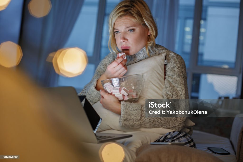 Pathetic girl observing sad film Portrait of lady with piteous face watching video on laptop. She is sitting and eating marshmallow. Copy space in left side Eating Stock Photo