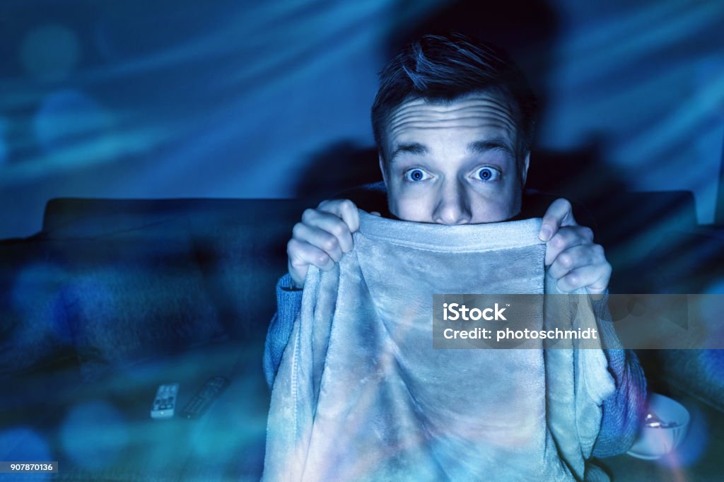 Man is afraid while watching tv A man is pulling up a blanket to cover his mouth while watching tv in a dark living room. Horror Movie Stock Photo