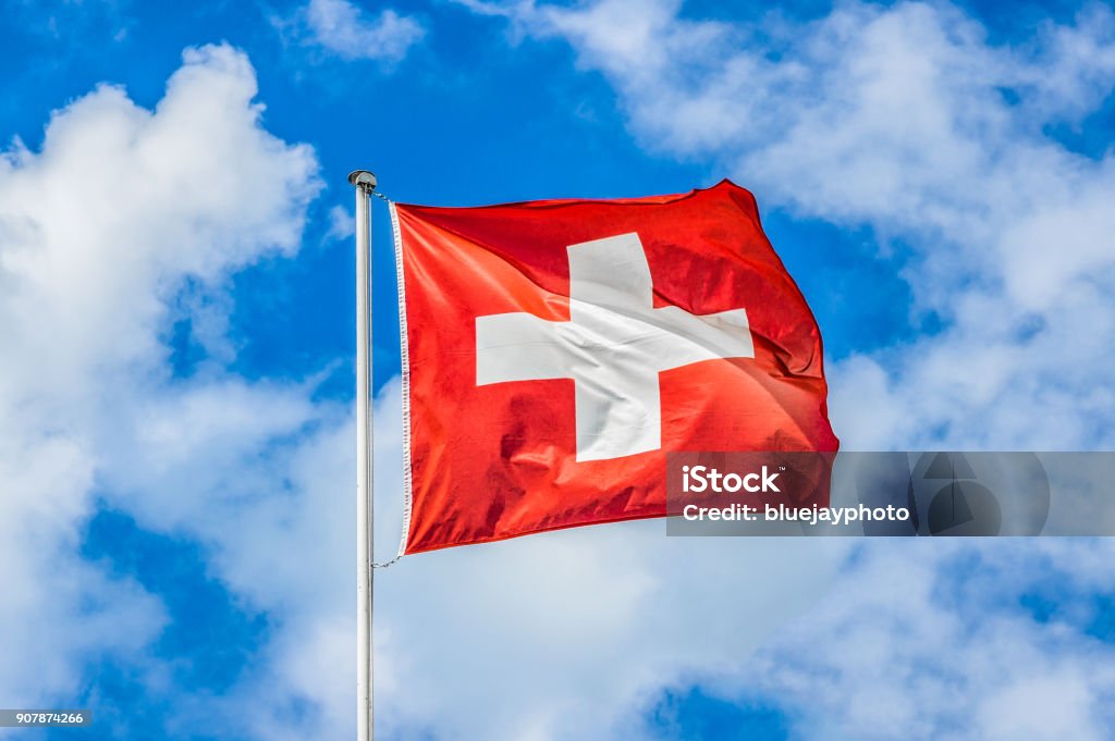 Swiss flag waving in the wind on a sunny day with blue sky and clouds Classic view of the national flag of Switzerland waving in the wind against blue sky and clouds on a sunny day in summer on the First of August, the national holiday of the Swiss Confederation Swiss Flag Stock Photo