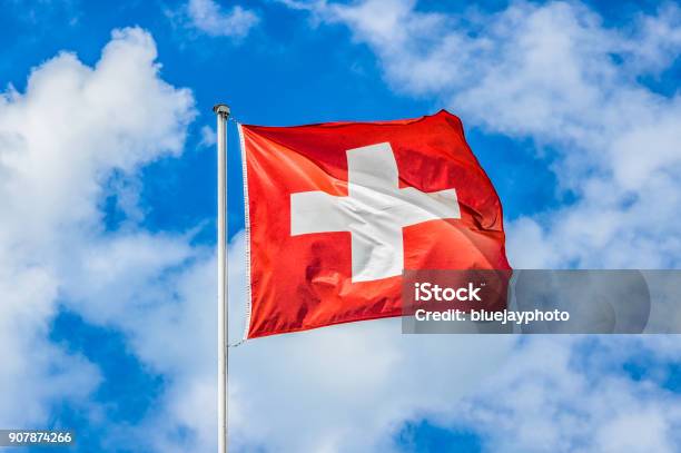 Swiss Flag Waving In The Wind On A Sunny Day With Blue Sky And Clouds Stock Photo - Download Image Now