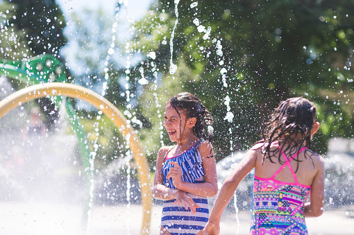 Two young girls in swimwear frolic and run in a playground splash pad. It is a sunny, perfect day for getting wet and playing hard! They are running through the sprinklers and yelling and laughing as the water droplets cascade all around them.