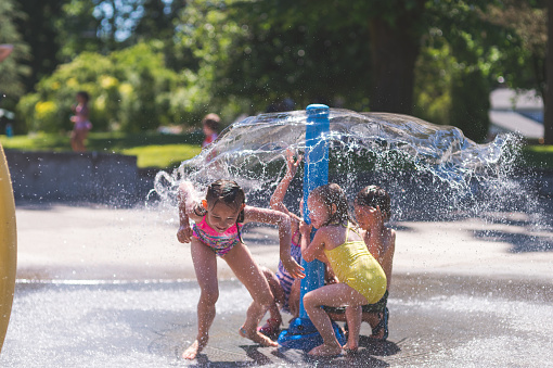 A group of of young children in swimwear duck and run around a sprinkler at a neighborhood splash pad. It is a sunny, perfect day for getting wet and playing hard! The water is coming out the top like an umbrella.