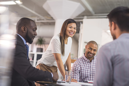 The CEO of a business startup gathers her multiethnic team around the conference table to finalize the launch. She is standing up and smiling as she listens to one of her colleagues pitch an idea.