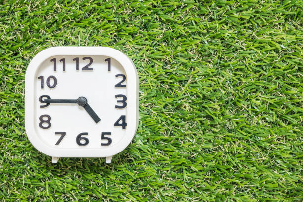 istock Closeup white clock for decorate show a quarter to five o'clock or 4:45 p.m. on green artificial grass floor textured background with copy space 907910578