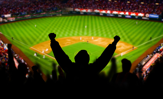A baseball fan raises his arms in celebration. The stadium is fictional.