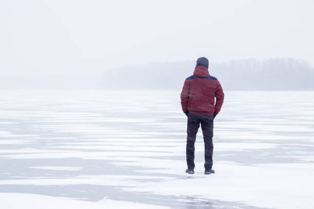 istock Young man in snow blizzard standing alone on the lake ice and staring forward. Cold atmosphere in the afternoon. Foggy air. Winter concept. 907927038