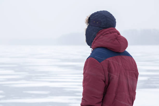 istock Young man in snow blizzard standing alone on the lake ice and staring forward. Cold atmosphere in the afternoon. Foggy air. Winter concept. 907927120