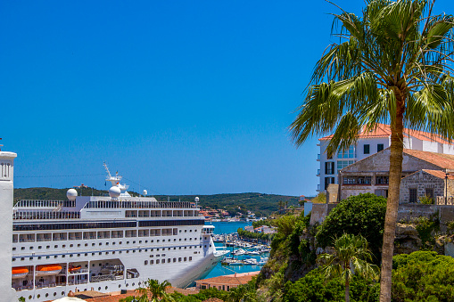 Spain, Menorca - June 13, 2014: A view of a white ship, yachts and boats standing at the pier in the port of Menorca. Green palm trees and trees with stone houses are a part of city life.