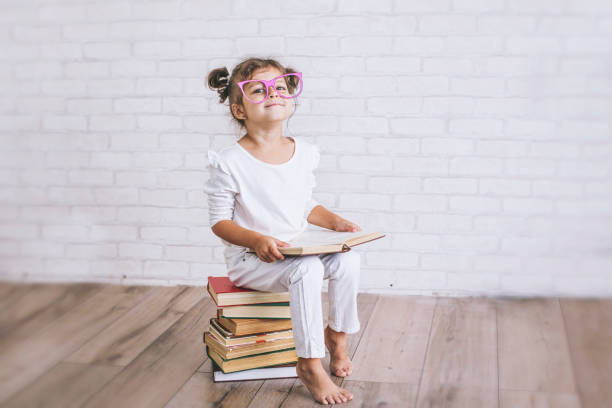 criança uma menina sentada sobre uma pilha de livros com óculos - child glasses elementary student reading - fotografias e filmes do acervo