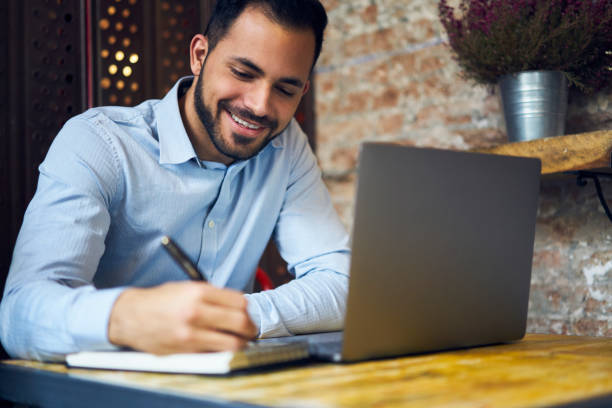 istock Cheerful handsome male designer drawing sketch in notepad before modeling on laptop computer application, young professional business manager making planning of startup sitting in cafe interior 907937298