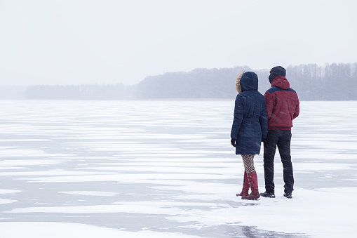 Young couple in snow blizzard walking on the lake ice and staring forward. Cold atmosphere in the afternoon. Foggy air. Winter concept.