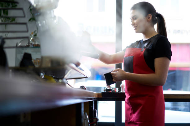 istock Attractive brunette female barista enjoying working process preparing coffee drinks using professional equipment in cafeteria.Skilled waitress in apron steaming milk on machine making cappuccino 907941882