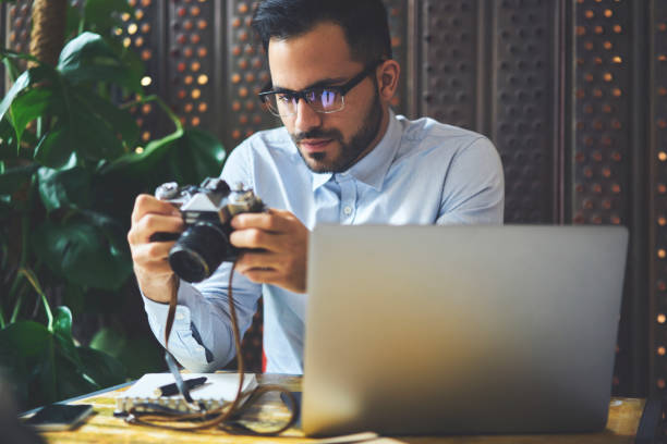 istock Pensive young male freelancer checking settings on vintage cameras sitting in cafe interior, skilled photographer testing usability of equipment for taking pictures and sharing multimedia on website 907941934