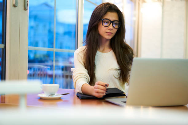 istock Pensive young brunette employee making researches in network watching videos analyzing information, professional project manager in eyewear working on laptop application having coffee break in cafe 907942010