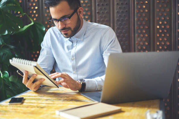 istock Handsome male entrepreneur checking working schedule in notepad while doing remote job in coworking space, pensive bearded designer proofreading strategy planning thinking about creation project 907942374