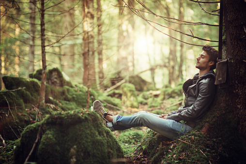 Young attractive man sitting in the forest and resting with eyes closed, relaxation and healthy lifestyle concept